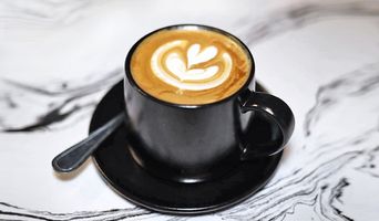 A latte in a black cup with saucer on a marble counter.