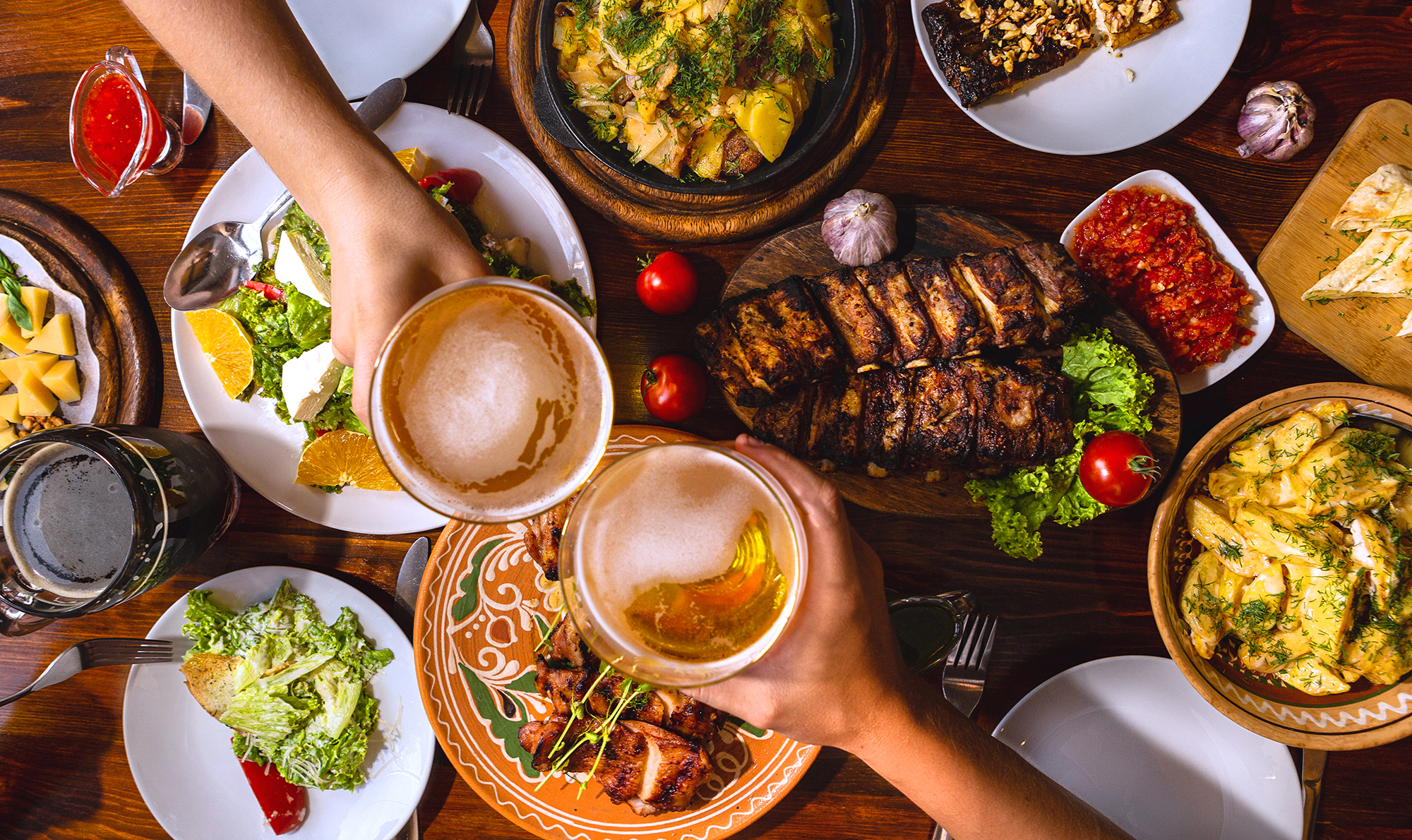 Two hands raise beers over a table with plated steaks and sides.