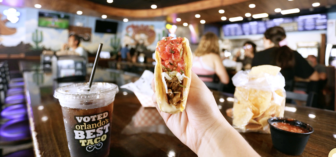 A hand holing a taco in a restaurant with people in the background.